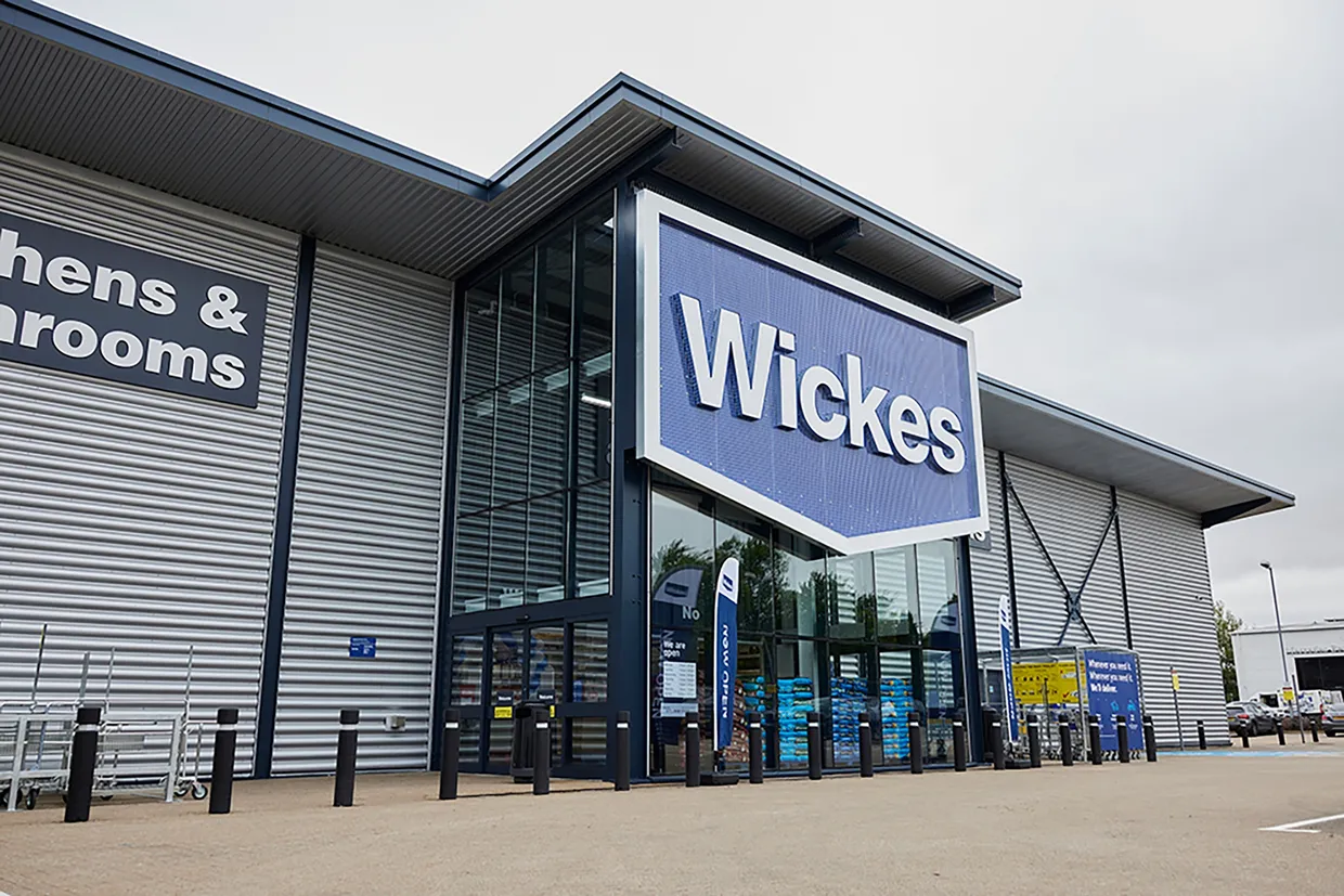 The exterior of the entrance of a Wickes' DIY store, with the large blue Wickes logo hanging over the glass-walled entrance